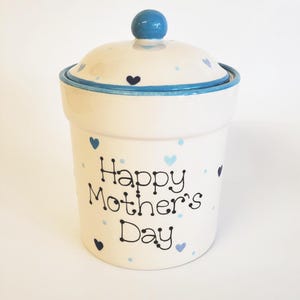 Könnte beinhalten: Weiße Keramikdose mit blauem Deckel und blauen und schwarzen Herz-Akzenten. Die Dose trägt den Schriftzug "Happy Mother's Day".