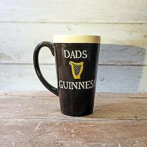 May include: Black ceramic mug with a white rim and the text "DADS GUINNESS" in white lettering with a gold harp logo.