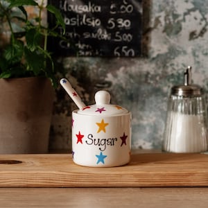 Peut inclure: Pot en céramique blanche pour le sucre avec un couvercle et une cuillère. Le pot est décoré d'étoiles colorées et du mot "Sugar" en lettres noires.