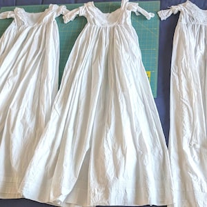 May include: Three white cotton gowns with lace trim and ruffled sleeves. The gowns are laid out on a green cutting mat.