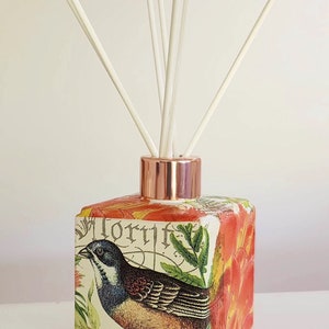 May include: A square glass diffuser bottle with a copper lid and white reeds. The bottle features a floral design with a bird and the word "Florif" printed on it.
