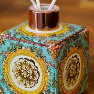 May include: Square ceramic diffuser bottle with a rose gold lid and floral pattern. The bottle has a blue background with gold accents and a floral design. Three white diffuser sticks are in the bottle.