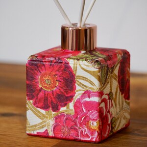 May include: A square glass diffuser bottle with a rose gold lid and floral design. The bottle has a floral pattern with pink and red flowers on a gold background. The bottle is filled with a clear liquid and has reeds sticking out of the top.