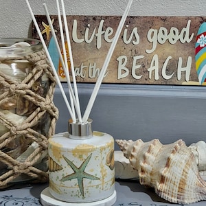 May include: A white diffuser bottle with a starfish design and a wooden sign that reads "Life is good at the beach" with a surfboard graphic.