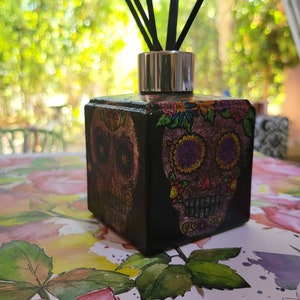 May include: A black square diffuser bottle with a silver lid and black reeds. The bottle is decorated with a colourful sugar skull design.