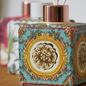 May include: A teal-coloured reed diffuser with a floral pattern and gold-coloured frame. The square container has a copper-coloured cap. The container's design features a central flower. Other diffusers are in the background.