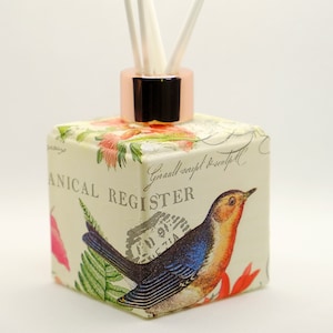 May include: A square glass diffuser bottle with a copper coloured lid and a floral design featuring a blue bird. The bottle has a label that reads "Botanical Register" and "Girall script! Beautiful".