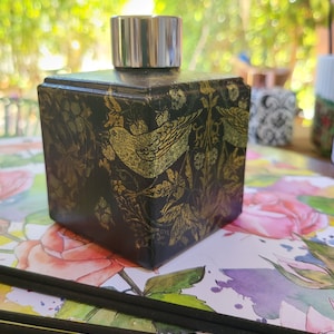 May include: A black square bottle with a silver lid and a gold floral design featuring birds. The bottle is sitting on a surface with a floral pattern.