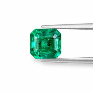May include: A vibrant green emerald gemstone with an emerald cut, featuring a rectangular shape with faceted edges. The gemstone is held by a pair of silver-toned metal tweezers, showcasing its translucent quality and deep green color saturation.