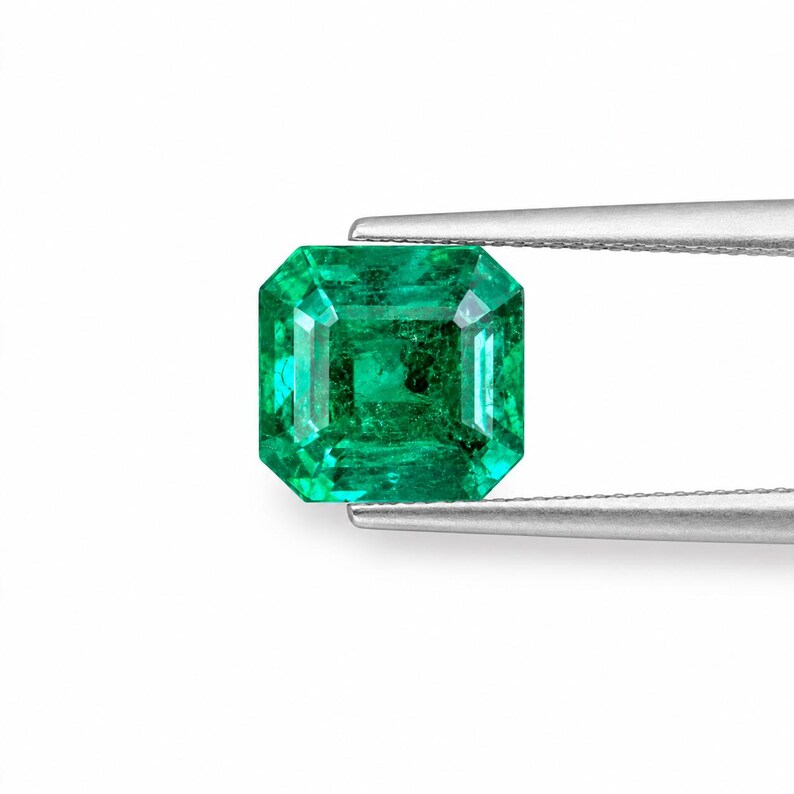 May include: A vibrant green emerald gemstone with an emerald cut, featuring a rectangular shape with faceted edges. The gemstone is held by a pair of silver-toned metal tweezers, showcasing its translucent quality and deep green color saturation.
