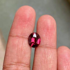 May include: A faceted, oval-shaped, deep red gemstone. The gem is held between two fingers, showcasing its polished surface and internal reflections. The stone's color ranges from a vibrant ruby to a darker, more subdued shade of red.