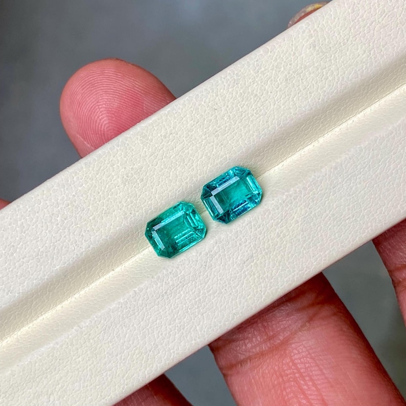 May include: Two rectangular, emerald-cut gemstones with a vibrant teal color. The gems are displayed on a white surface, showcasing their facets and clarity. The stones are held in a hand, highlighting their size and shape.