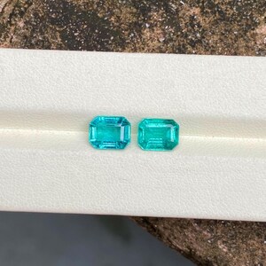 May include: Two rectangular, teal-colored gemstones with a step-cut facet pattern. The gems are displayed on a white surface, with a textured, brown background. The gems are likely for jewelry making or collecting.