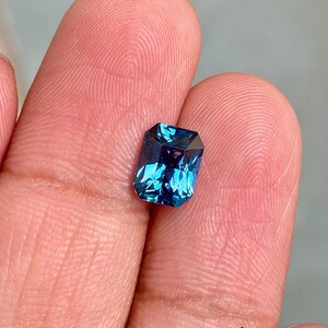 May include: A deep blue gemstone with a rectangular emerald cut. The gemstone is held in a hand.