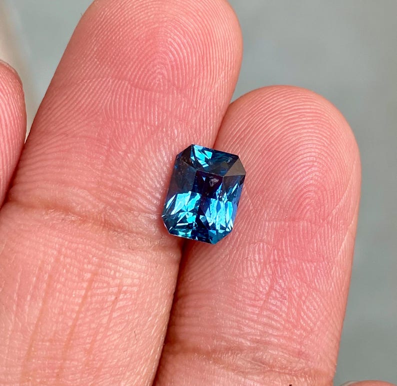 May include: A deep blue gemstone with a rectangular emerald cut. The gemstone is held in a hand.