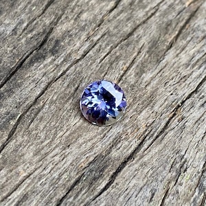 May include: A round, faceted, purple gemstone with a slight blue hue. The gemstone is set against a rustic wooden background.
