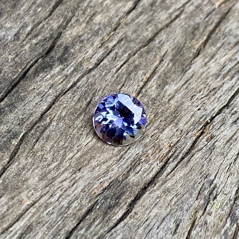 May include: A round, faceted, purple gemstone with a slight blue hue. The gemstone is set against a rustic wooden background.
