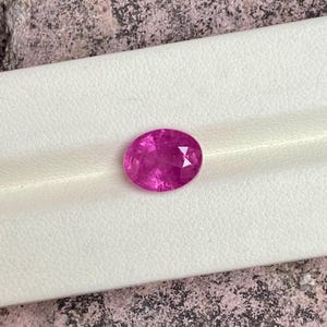 May include: A faceted, oval-shaped, hot pink gemstone. The gem is set against a white background. The stone has a brilliant cut, reflecting light. The colour is a vibrant, saturated pink, with a high degree of clarity.