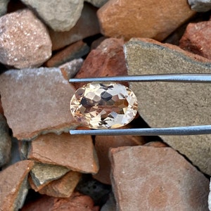 May include: A light brown oval-shaped gemstone with a faceted cut. The gemstone is held by tweezers against a background of brown rocks.