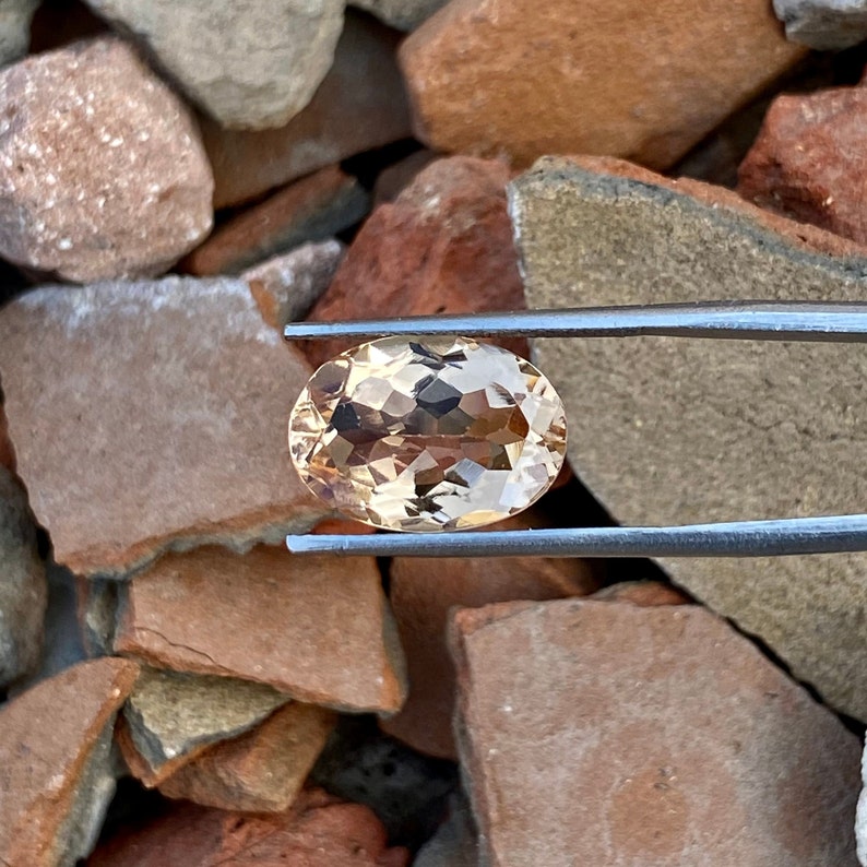 May include: A light brown oval-shaped gemstone with a faceted cut. The gemstone is held by tweezers against a background of brown rocks.