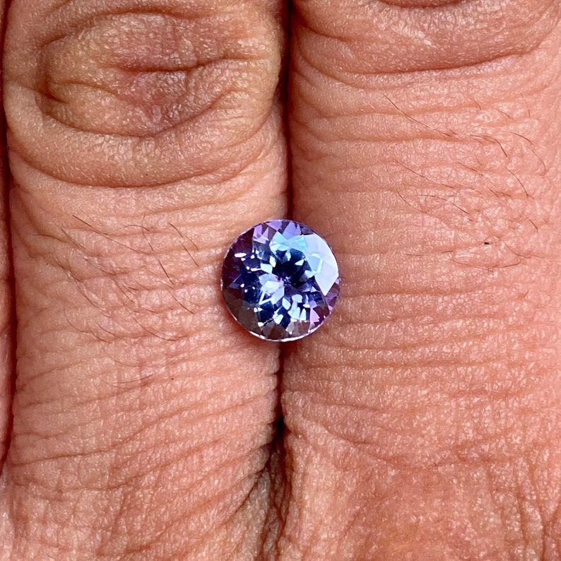 May include: A round, faceted gemstone with a purple and blue colour gradient. The gemstone is set in a ring and is being held by a hand.