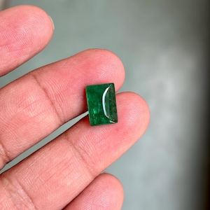 May include: A rectangular, emerald-green gemstone with a polished surface. The gem is held between fingers, showcasing its vibrant color and clarity. The stone's shape is a rectangle with slightly rounded corners.