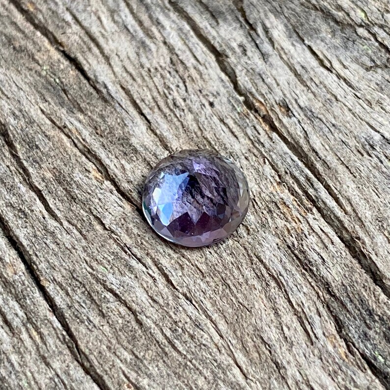 May include: A round, faceted gemstone with a purple and blue iridescent sheen. The gemstone is set against a rustic wooden background.