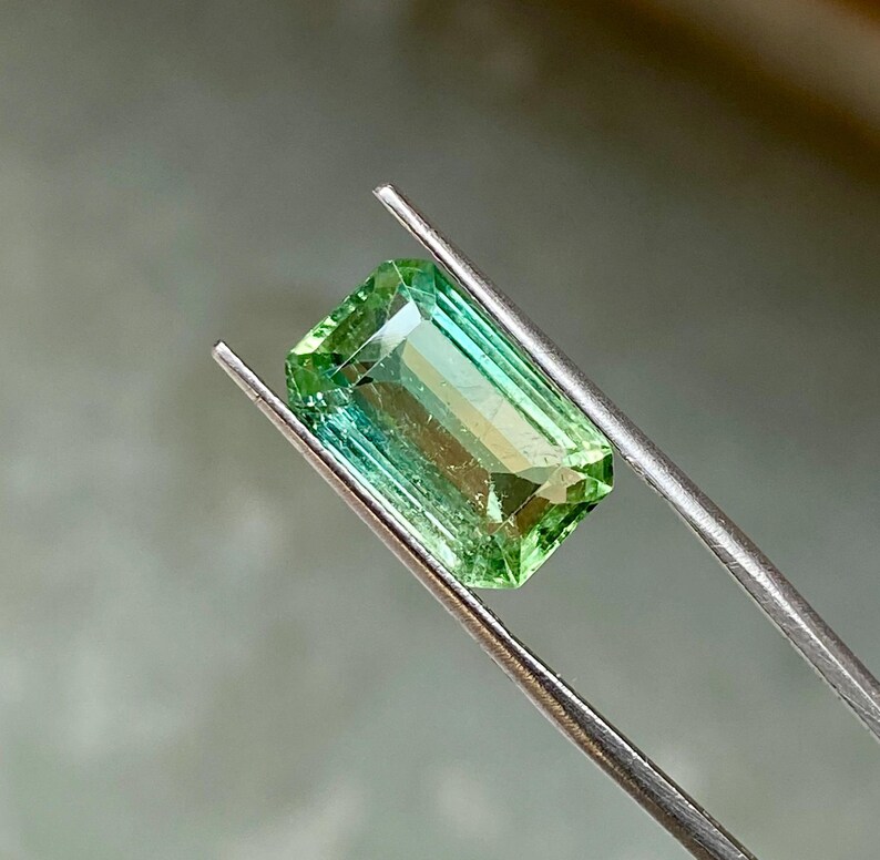 May include: A green gemstone with a rectangular emerald cut, held by tweezers. The gemstone has a light green color at the top and a darker green color at the bottom.