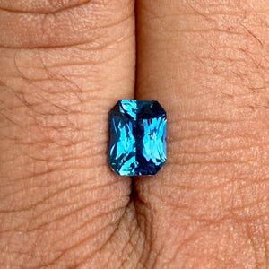 May include: A deep blue gemstone with a rectangular emerald cut. The gemstone is set in a ring and is surrounded by a silver band.