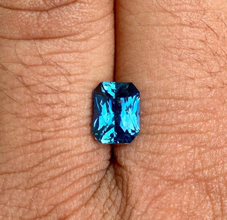 May include: A deep blue gemstone with a rectangular emerald cut. The gemstone is set in a ring and is surrounded by a silver band.