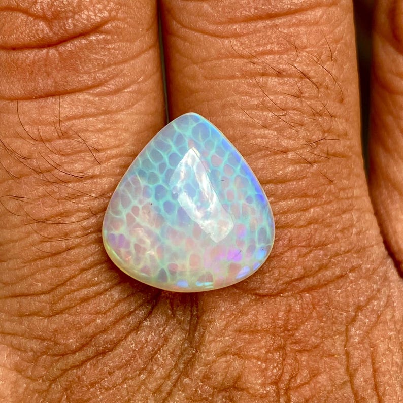 May include: A teardrop-shaped opal gemstone with a white, blue, and iridescent finish. The opal has a unique pattern of small, white dots.