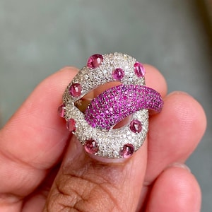 May include: A unique ring in the shape of a mouth, featuring a tongue and lips. The lips are encrusted with small, sparkling white stones, while the tongue is covered in smaller, vibrant pink stones. Pink gemstones adorn the outer edges.