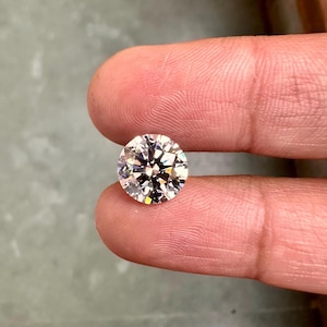 May include: A clear round diamond gemstone with facets, held between two fingers.