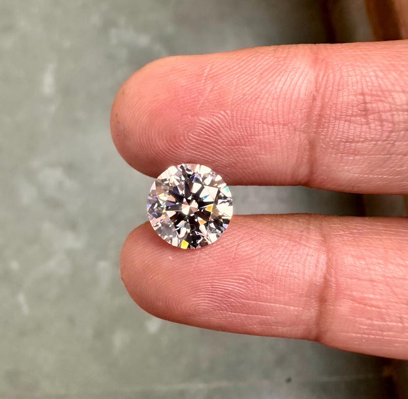 May include: A clear round diamond gemstone with facets, held between two fingers.