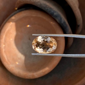 May include: A light brown oval-shaped gemstone held by tweezers. The gemstone is faceted and has a translucent appearance.