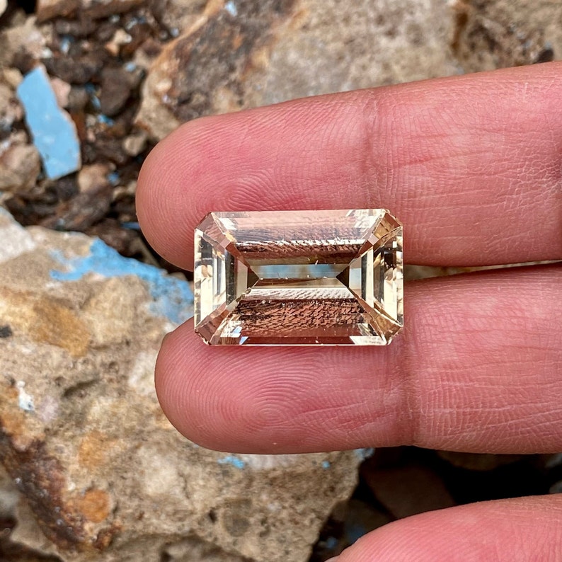 May include: A light brown, faceted, rectangular gemstone with a step cut. The gemstone is held in a hand.