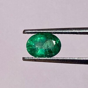May include: A vibrant green emerald gemstone, oval-shaped, held by tweezers against a light pink background.