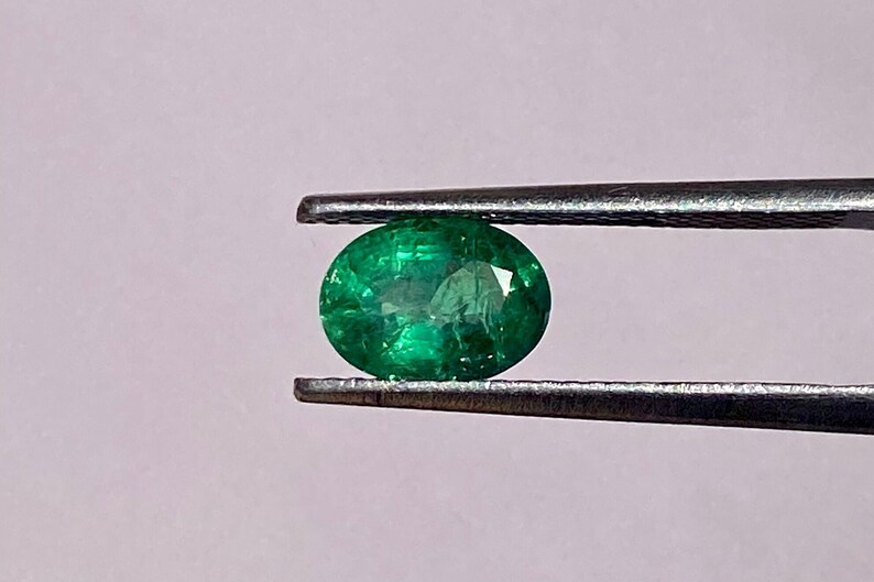 May include: A vibrant green emerald gemstone, oval-shaped, held by tweezers against a light pink background.