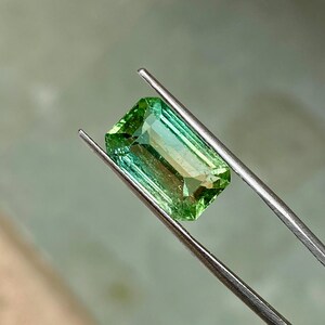 May include: A green gemstone with a rectangular emerald cut. The gemstone is held in tweezers.