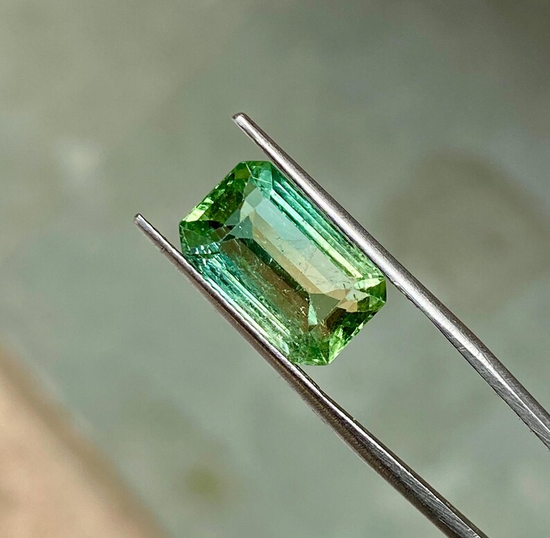 May include: A green gemstone with a rectangular emerald cut. The gemstone is held in tweezers.