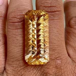 Puede incluir: Una gran gema de citrino dorado facetada con forma rectangular. La gema está tallada con un corte de escalón y tiene un brillo brillante.