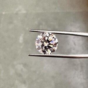 May include: A clear round diamond gemstone held by tweezers against a grey background.