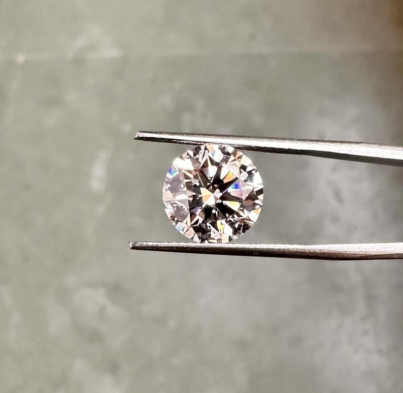 May include: A clear round diamond gemstone held by tweezers against a grey background.