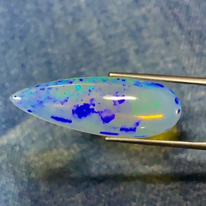 May include: A teardrop-shaped opal gemstone with a white base and blue, green, and purple iridescent flashes. The opal is held in tweezers.