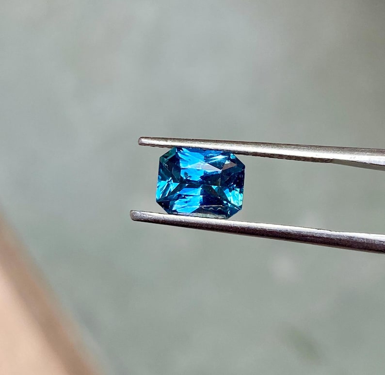 May include: A vibrant blue gemstone with a rectangular emerald cut, held by tweezers.
