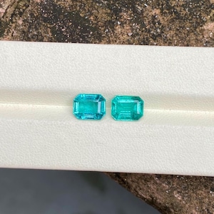 May include: Two rectangular, teal-colored gemstones with a step-cut design. The gems are displayed on a white surface, showcasing their clarity and vibrant color. The gems are likely for jewelry making.