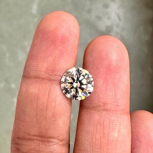 May include: A clear round diamond gemstone with facets, held between two fingers.