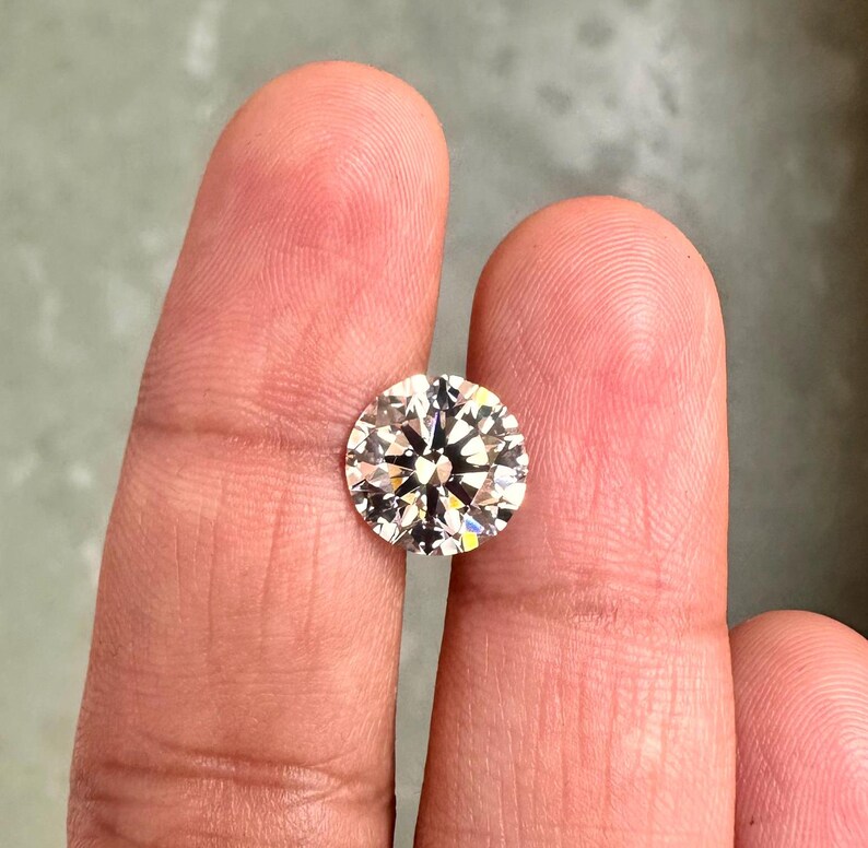 May include: A clear round diamond gemstone with facets, held between two fingers.