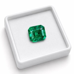 May include: A vibrant, emerald-green gemstone with a rectangular step-cut, featuring distinct facets that catch the light. The loose stone is centered within a square, white display box that has a soft, padded interior lining for protection.