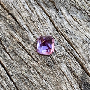 Puede incluir: Una piedra preciosa facetada de forma cuadrada con un tono p&uacute;rpura y rosa. La piedra preciosa est&aacute; colocada sobre un fondo de madera.